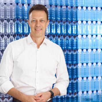 A man in a white business shirt smiles at the camera in front of a background of blue plastic bottles
