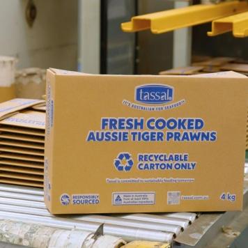 A brown cardboard box with blue writing on it sits on a conveyor belt with stacks of flattened boxes behind it