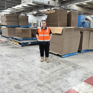 A woman wearing an orange high vis safety top smiles at camera, behind her is a concrete area with stacks of brown cardboard