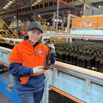 A woman with black cap and safety glasses smiles at camera holding a green wine bottle. She is in a factory with lots of green wine bottles behind her on a conveyor belt.