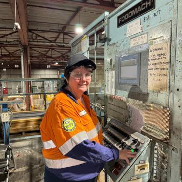 A woman smiles at camera in a black cap, orange and blue high vis safety work wear standing in a factory