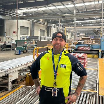 Man in high-vis top and baseball cap stands and smiles at the camera, with cardboard box production behind him.