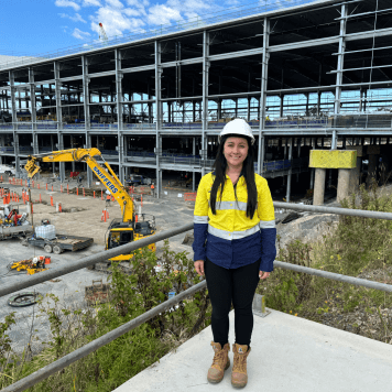 Woman in high-vis top and safety helmet smiles while standing in front of construction site.