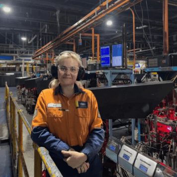 Woman in high-vis top and safety helmet smiles while standing in front of glass machinery.