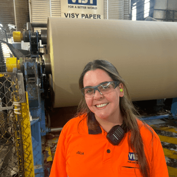 Woman in orange high-vis top smiles at the camera in front of a paper machine.