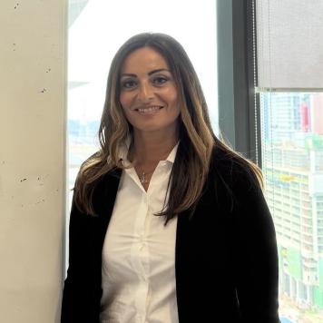 Woman in white shirt and black cardigan smiles at the camera, standing in front of a window.