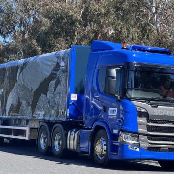 A blue Visy Logistics truck trailer driving on a road with large green gum trees behind it