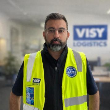 Man wearing yellow high-vis vest and glasses smiles at the camera, in a Visy office.