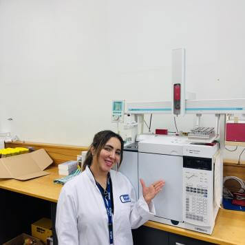 Woman with dark hair, wearing a white lab coat, smiles at her desk.