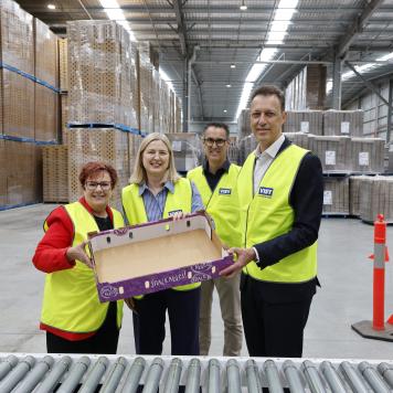 Four people in high visibility safety vests are holding a cardboard produce tray smiling at camera, standing in a warehouse