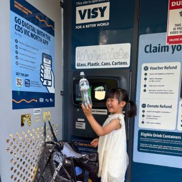 A little girl in a white dress holds up a plastic bottle in front of a reverse vending machine