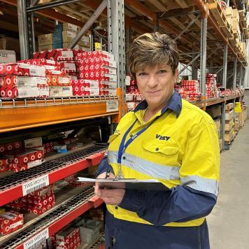 A woman smiles at camera. She is wearing high visibility, safety work wear and holding a clipboard, standing in a warehouse with lots of items on shelves