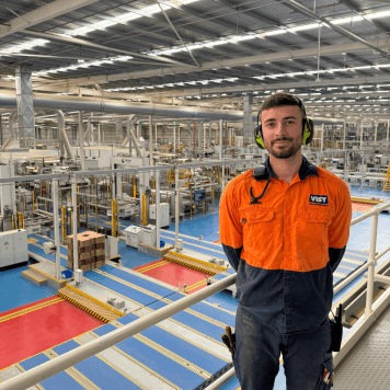 Jarrod, a Visy electrician, is wearing a high-vis orange shirt. He is smiling at the camera in front of a cardboard box facility.