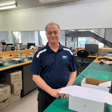 Claudio, a Visy Lab Tech, wears a navy polo shirt with the Visy logo. He stands in a laboratory and smiles.