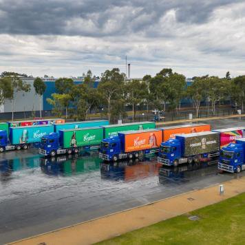 6 Visy Logistics trucks lined up on wet tarmac with brightly coloured Coopers Brewery livery, trees and warehouses in the background