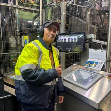 Rebecca wears PPE and smiles at the camera with a thumbs-up. She is standing next to beverage can making equipment.