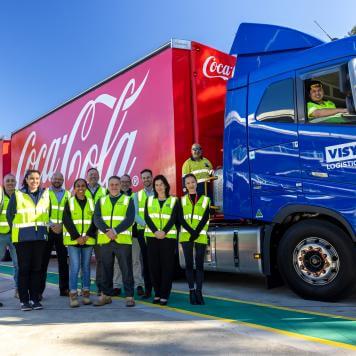 14 people in high visibility safety vests stand in front of a red Coca Cola truck with a blue Visy Logistics front