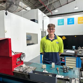 Max wears high-vis and PPE, and smiles at the camera while working in the tool shop