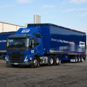 Two blue trucks with Driven by Visy Global Logistics on them in front of a warehouse