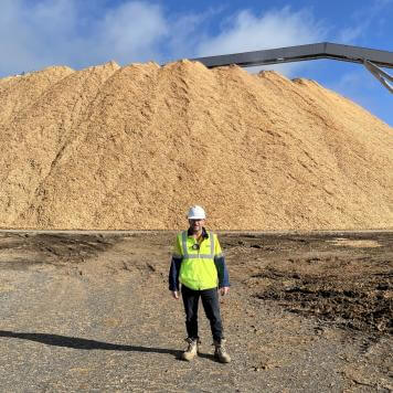 Brad wears PPE and high-vis vest, and stands and smiles in front of a large pile of wood bark..